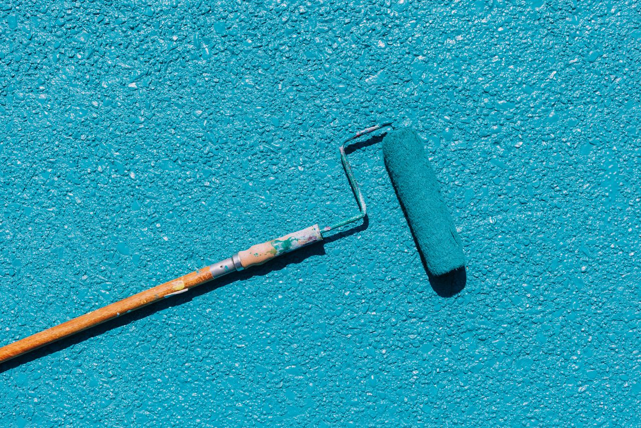 A close-up of a blue paint roller applying paint on a textured wall surface, captured in bright sunlight.